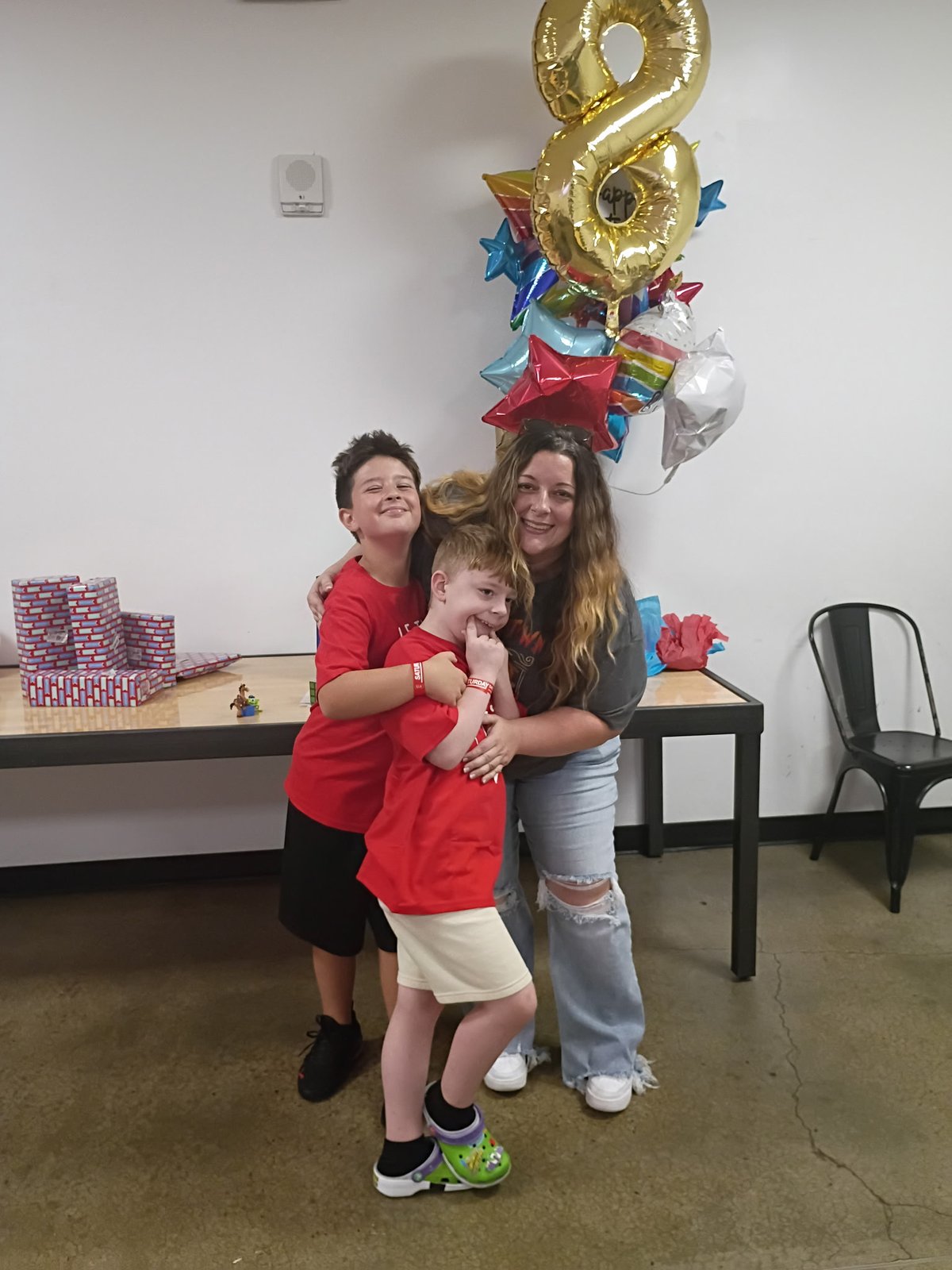 Nicole smiling with her arms around Connor and his brother, both in red shirts, at a birthday celebration with a gold number 8 balloon and star balloons behind them.