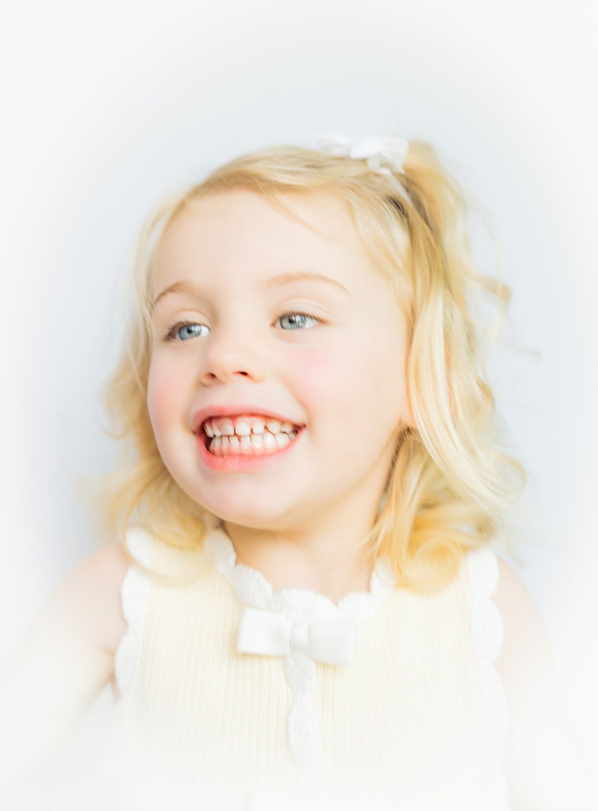 Norah, a young child with blonde hair and a white bow, smiling in a pale yellow sleeveless top.