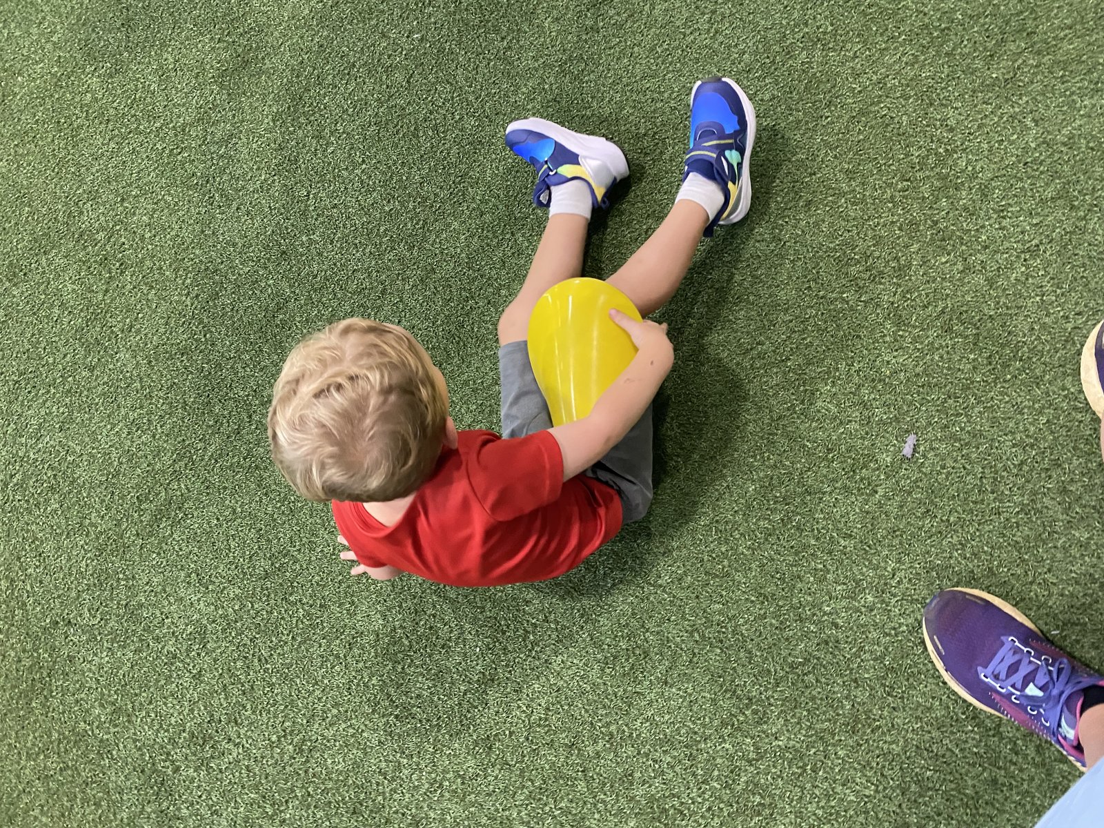Young boy in a red shirt sitting on green turf holding a yellow sports cone (overhead view at soccer/practice).