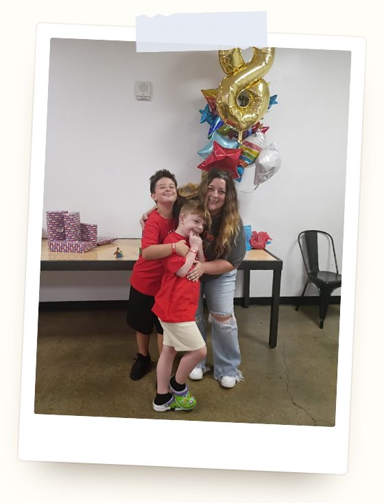 Nicole smiling with her arms around Connor and his brother, both in red shirts, at a birthday celebration with a gold number 8 balloon and star balloons behind them.